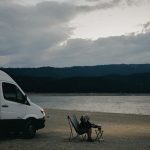 A woman relaxes by a lakeside, seated next to a campervan with a dog and folding chair under cloudy skies