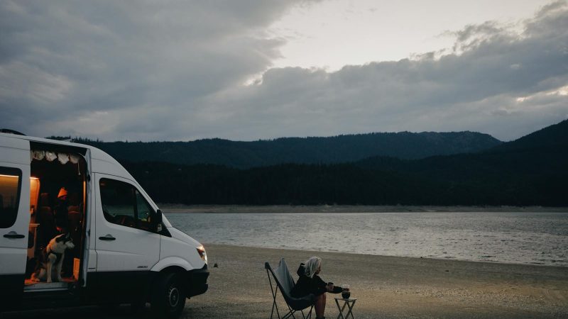 A woman relaxes by a lakeside, seated next to a campervan with a dog and folding chair under cloudy skies